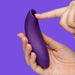 A hand holds the We-Vibe Melt 2 Rechargeable Pleasure Air Clitoral Stimulator as another hand points to its round opening, highlighting its waterproof, smooth silicone design against a solid purple background.