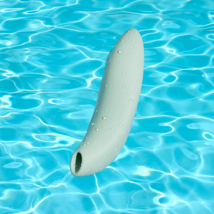 A pale green We-Vibe Melt 2 Rechargeable Pleasure Air Clitoral Stimulator with a waterproof body and water droplets floats upright in a clear, blue swimming pool.