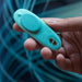 A hand holds the We-Vibe Moxie +, a small turquoise panty vibrator with two gold buttons and an oval area. The discreet wearable can be controlled via remote or app. Blue abstract lines form the background.