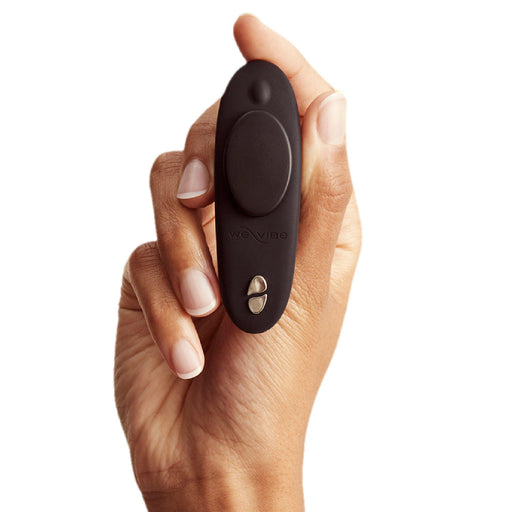 A hand holds the We-Vibe Moxie +, a small, black, oval-shaped panty vibrator with buttons, designed for discreet hands-free use and remote or app control, against a white background.