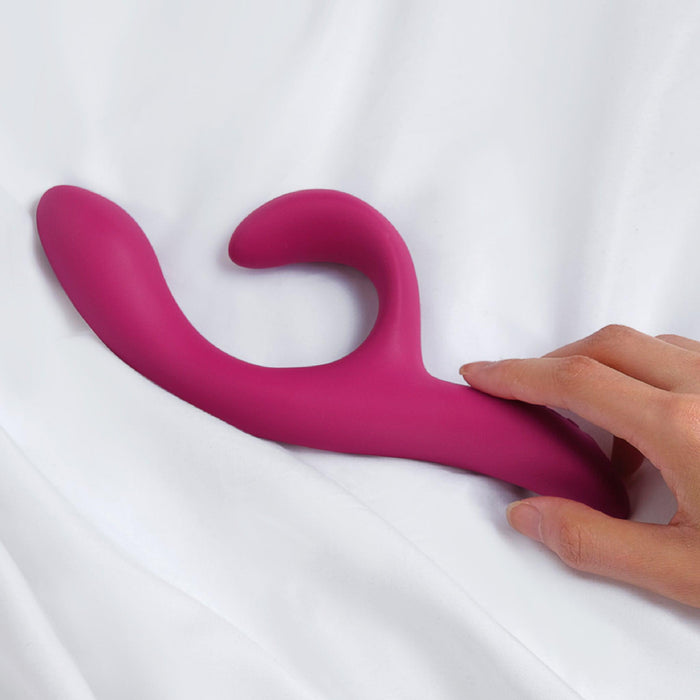 A hand reaches for the We-Vibe Nova 2 Powerful Flexible G-Spot Rabbit Vibrator, pink and curved, resting on white bed sheets.