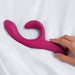 A hand reaches for the We-Vibe Nova 2 Powerful Flexible G-Spot Rabbit Vibrator, pink and curved, resting on white bed sheets.