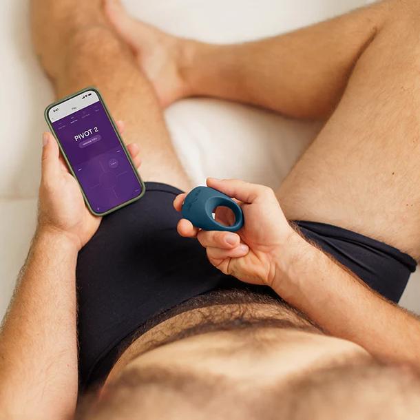 A person in black underwear lies on a bed, holding a smartphone displaying customizable vibration patterns and the We-Vibe Pivot 2 Stretchy Vibrating Silicone Rechargeable Penis Ring for Couples.