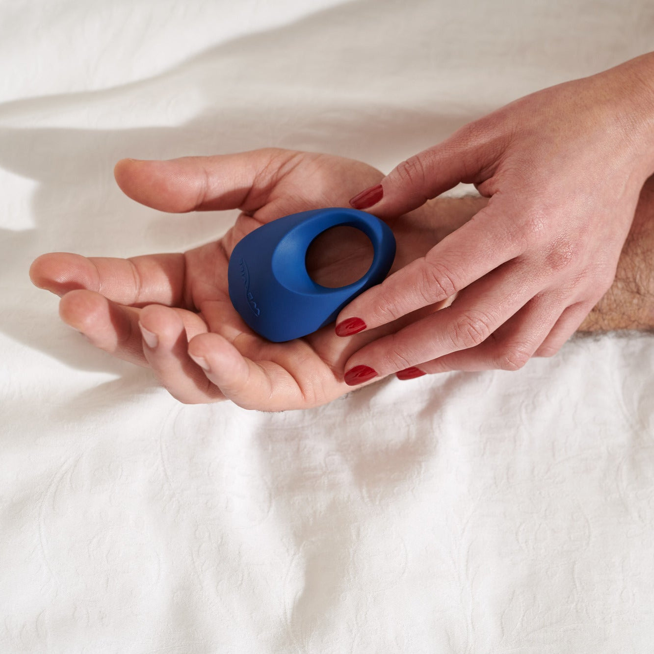 A person with red nail polish places a blue We-Vibe Pivot Vibrating Silicone Rechargeable Penis Ring with App Control into another’s open palm, both hands resting on a white bedsheet.