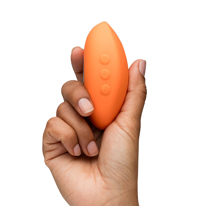 A hand holds the orange, oval-shaped We-Vibe Temp Lay On Vibrator with hot and cold functions and three raised buttons, against a white background.