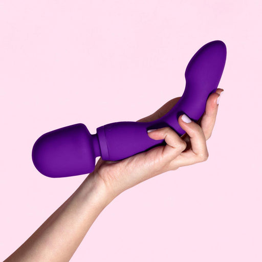 A hand with manicured nails holds the Blush Wellness Dual Sense Double Ended Ergonomic Waterproof Wand, a two-part purple g-spot vibrator, against a light pink background. The toy features a curved design with a bulbous end and a wand-like handle.