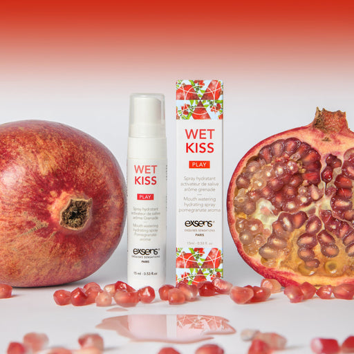 A bottle and box of Wet Kiss Pomegranate Moisturizing Saliva Spray are shown next to a whole pomegranate, a halved pomegranate, and scattered seeds on a white surface with a red gradient background.