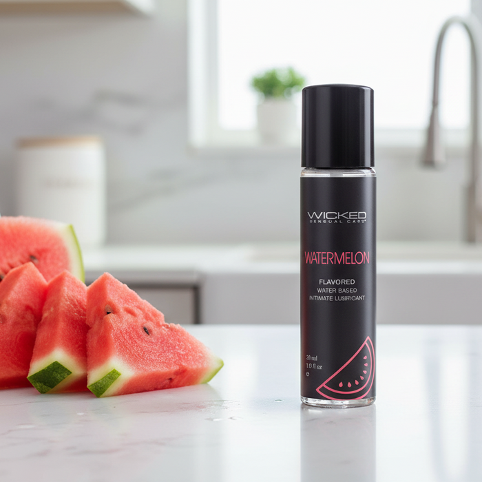 A 1 oz bottle of Wicked Aqua Mini Watermelon Flavored Water Based Lubricant rests on a kitchen counter beside sliced watermelon, with the background softly blurred.