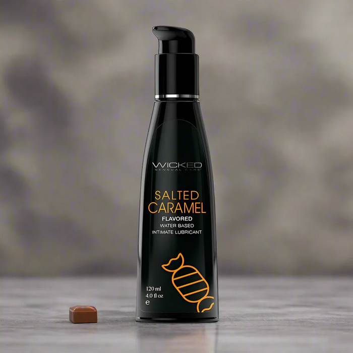 A 4 oz black bottle of Wicked Aqua Salted Caramel Flavored Water Based Lubricant stands upright on a gray surface with a caramel candy, set against a blurred gray background. This lubricant is sugar-free and deliciously smooth.