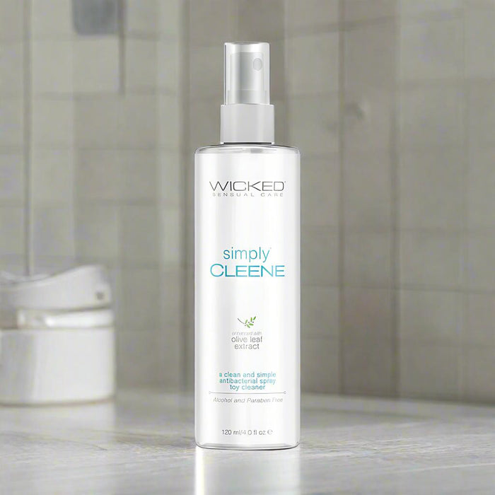 A clear 4 oz spray bottle labeled "Wicked Simply Cleene Antibacterial Toy Cleaner" sits on a bathroom counter. Alcohol-free and made with natural olive leaf extract, it offers a gentle yet effective clean for toys.