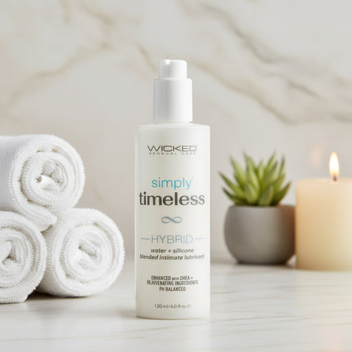 A bottle of Wicked Simply Timeless Hybrid Long Lasting Lubricant - 4 oz sits on a counter beside rolled white towels, a small potted succulent, and a lit candle, set against a marble background.