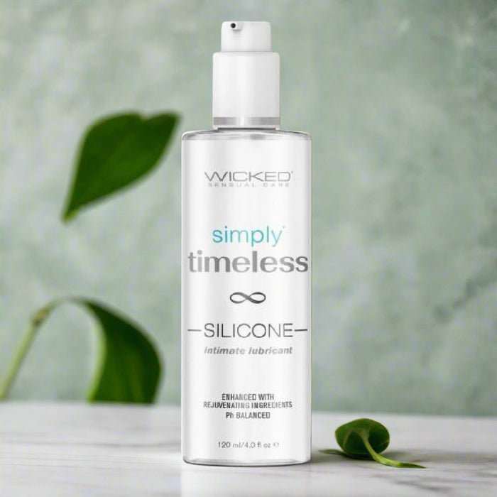 A 4 oz bottle of Wicked Simply Timeless Silicone Lubricant with Vitamin E, featuring a pump top, sits on a white surface with green leaves and a soft green background, highlighting its long-lasting glide and moisturizing formula.