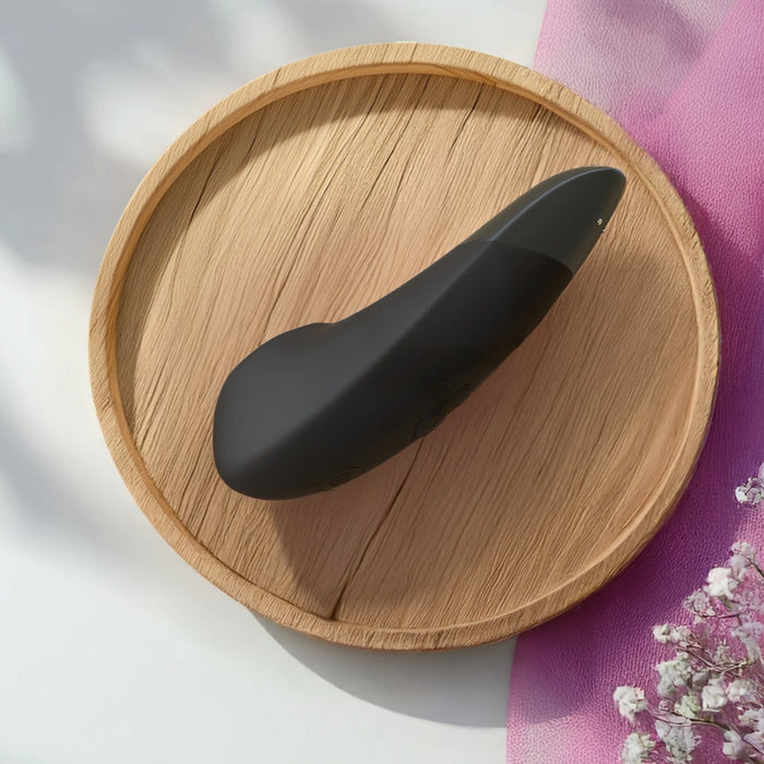 The Womanizer Enhance Pleasure Air + Vibration Clit Stimulator, a black curved device with rumbly vibrations, rests on a round wooden tray in sunlight near pink flowers and a pink cloth in the bottom right corner.