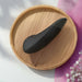 The Womanizer Enhance Pleasure Air + Vibration Clit Stimulator, a black curved device with rumbly vibrations, rests on a round wooden tray in sunlight near pink flowers and a pink cloth in the bottom right corner.