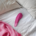 The Womanizer Enhance Pleasure Air + Vibration Clit Stimulator in pink rests on a white bedspread beside a pink blanket and scarf.