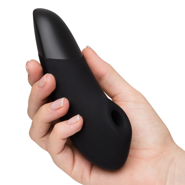 A hand holds the Womanizer Enhance Pleasure Air + Vibration Clit Stimulator, a sleek black silicone device with a rounded tip and circular opening near the base, featuring Pleasure Air Technology, against a plain white background.