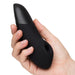 A hand holds the Womanizer Enhance Pleasure Air + Vibration Clit Stimulator, a sleek black silicone device with a rounded tip and circular opening near the base, featuring Pleasure Air Technology, against a plain white background.
