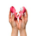 Two hands hold the Womanizer Liberty 2 Pleasure Air Travel Sized Clitoral Stimulator, a pink silicone vibrator with white accents and buttons, featuring a curved design for clitoral pleasure via air technology against a plain white background.