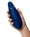 A hand holds a dark blue Womanizer Premium 2 Pleasure Air Clitoral Stimulator, featuring three raised buttons and a metallic accent near the top, shown against a white background.
