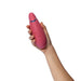A hand holds the Womanizer Premium 2 Pleasure Air Clitoral Stimulator, a pink, waterproof, oval device with side buttons and a metallic ring near the top—designed for comfort and easy use with gentle air pulsation technology.