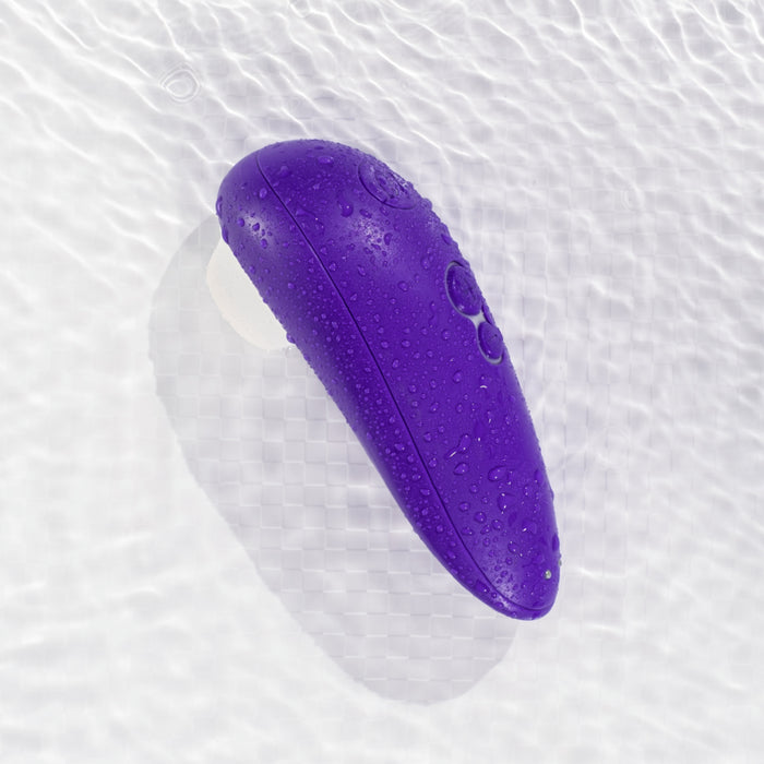 The Womanizer Starlet 3 Pleasure Air Clitoral Stimulator, a purple, water-resistant, oval-shaped device with buttons, is displayed on a rippled white surface and covered in water droplets.