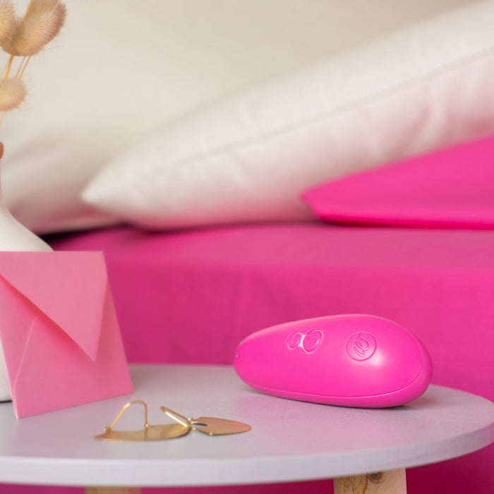 A pink Womanizer Starlet 3 Pleasure Air Clitoral Stimulator rests on a small round table with a pink envelope and gold jewelry, while a bed with pink sheets and white pillows appears in the background.