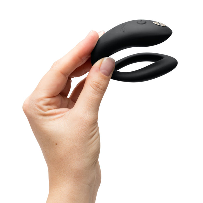 A hand holds the black, U-shaped Womanizer x We-Vibe Lust & Luxury Collection couples vibrator with control buttons, inspired by Pleasure Air technology, against a white background.