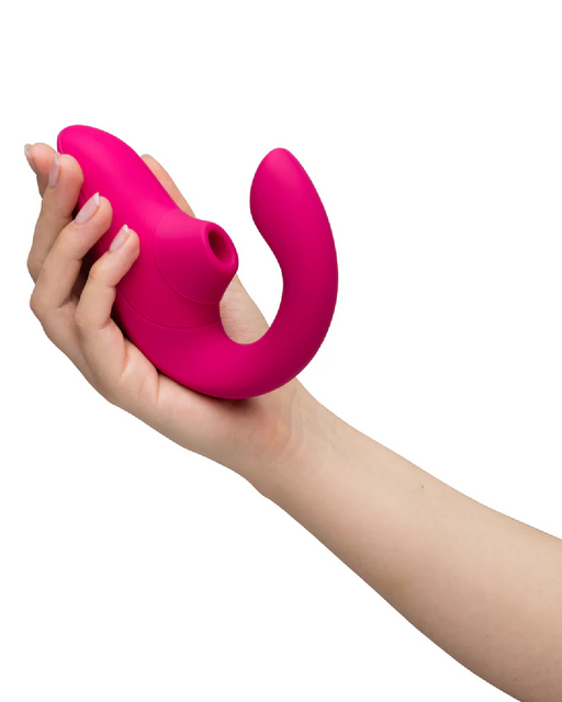 A hand is holding a bright pink, curved Womanizer Blend Pleasure Air Clitoral & G-Spot Rabbit - Pink toy designed for intimate use. The toy has a smooth, ergonomic shape and a rounded end for internal stimulation. The background is plain white, drawing attention to the toy and the hand holding it.
