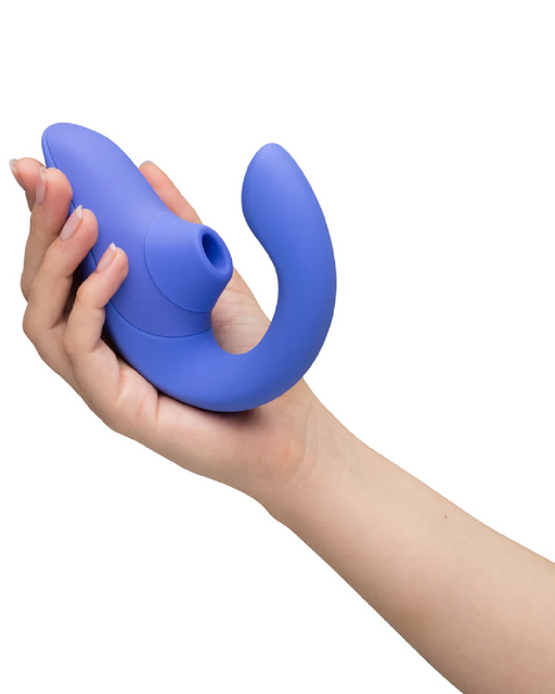A hand holding a sleek, blue Womanizer Blend Pleasure Air Clitoral & G-Spot Rabbit - Blue by Womanizer with a curved end and a small suction nozzle is featured. The ergonomically shaped adult toy, designed for dual stimulation, appears to be made of smooth, soft material against a plain white background.