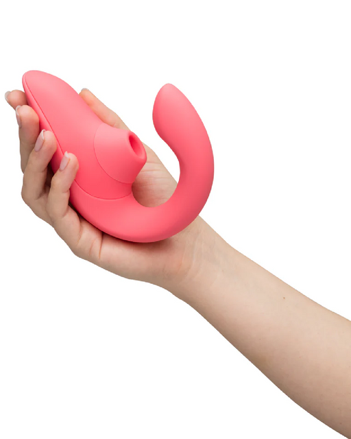 A hand is holding a pink, curved, silicone Womanizer Blend Pleasure Air Clitoral & G-Spot Rabbit - Rose adult toy designed for stimulation. The toy features a section for suction and a curved arm for internal use, providing dual stimulation. The background is white, putting full focus on the object.