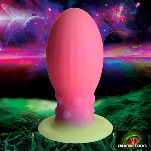 The Xeno Egg Glow In The Dark Extra Large Thick Silicone Egg, a large pink and green textured toy, is shown against a cosmic alien landscape with colorful planets and swirling clouds. The Creature Cocks logo is in the bottom right corner.