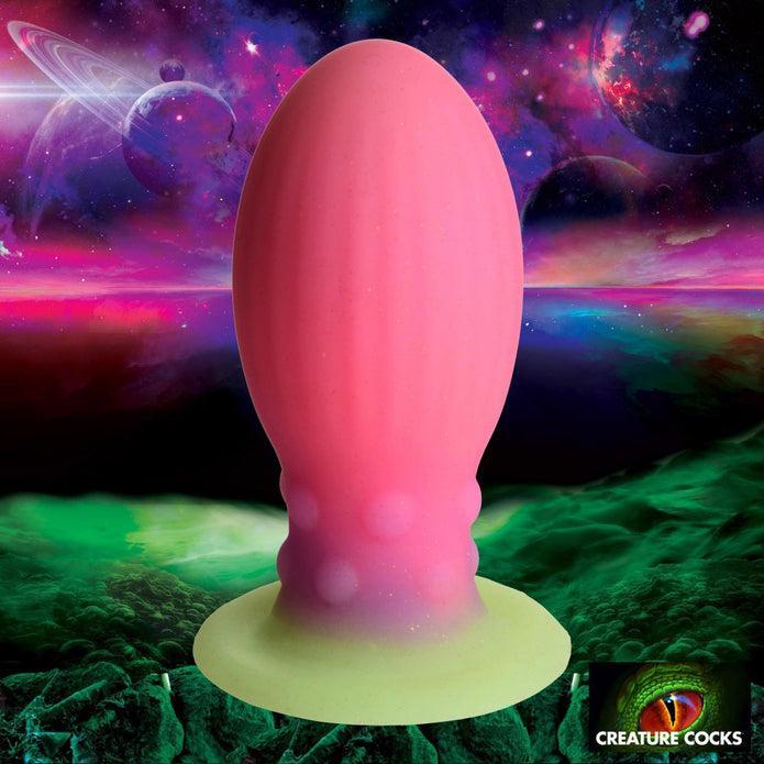 The Xeno Egg Glow In The Dark Extra Large Thick Silicone Egg, a large pink and green textured toy, is shown against a cosmic alien landscape with colorful planets and swirling clouds. The Creature Cocks logo is in the bottom right corner.