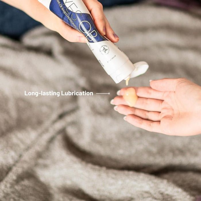 A person squirts beige AH! YES OB Plant-Oil Based Personal Lubricant, labeled "Long-lasting Lubrication," onto another's open palm against a background of gray fabric.