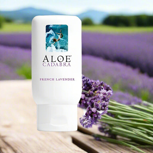 A bottle of Aloe Cadabra French Lavender Aloe Based Gentle Lubricant 2.5 oz rests on a wooden surface by fresh lavender, with a blurred lavender field and mountains behind, highlighting its organic aloe vera formula for pure relaxation and comfort.