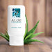 A 2.5 oz white bottle of Aloe Cadabra Natural Aloe Based Gentle Lubricant rests on a wooden surface with fresh aloe vera leaves beside it. The label features a woman swimming underwater, emphasizing its gentle, aloe-based formula.