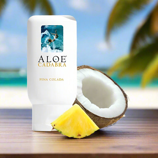 A bottle of Aloe Cadabra Pina Colada Aloe Based Gentle Lubricant 2.5 oz rests on a wooden surface beside a halved coconut and pineapple slice, with a blurred tropical beach and palm trees in the background.