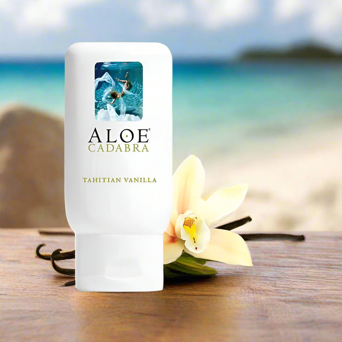 Aloe Cadabra Tahitian Vanilla Aloe Based Gentle Lubricant 2.5 oz rests on a wooden table next to a vanilla flower and bean, with a blurred view of a tropical beach and ocean in the background.