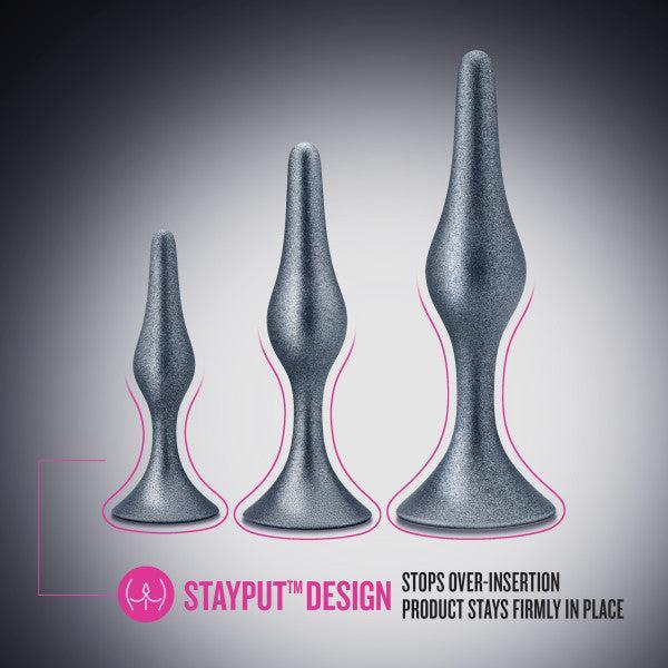 Three grey butt plugs of varying sizes from the Anal Adventures First Time Kit by Blush are presented vertically, with the smallest on the left and the largest on the right. A highlighted caption reads, "STAYPUT™ DESIGN" with text underneath stating, "Stops over-insertion; product stays firmly in place." Discover the innovative Anal Adventures First Time Kit.