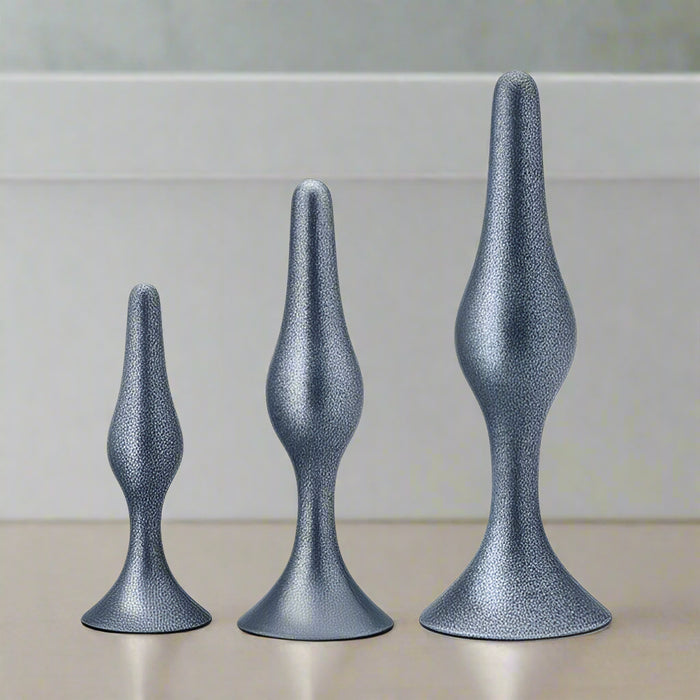 Three metallic, silver-colored butt plugs from the Blush Anal Adventures First Time Kit are aligned from smallest to largest against a plain white background. Each plug features a slightly textured, glittery finish and includes StayPut technology with a flared base for safety, making them perfect for anal training.