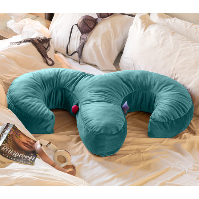 A teal Liberator Arie Toy Mount Snuggle Pillow with two vibrator pockets sits on an unmade bed with white and beige linens, surrounded by a bra, over-ear headphones, a book, and lipstick for a cozy, inviting vibe.