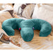 A teal Liberator Arie Toy Mount Snuggle Pillow with two vibrator pockets sits on an unmade bed with white and beige linens, surrounded by a bra, over-ear headphones, a book, and lipstick for a cozy, inviting vibe.
