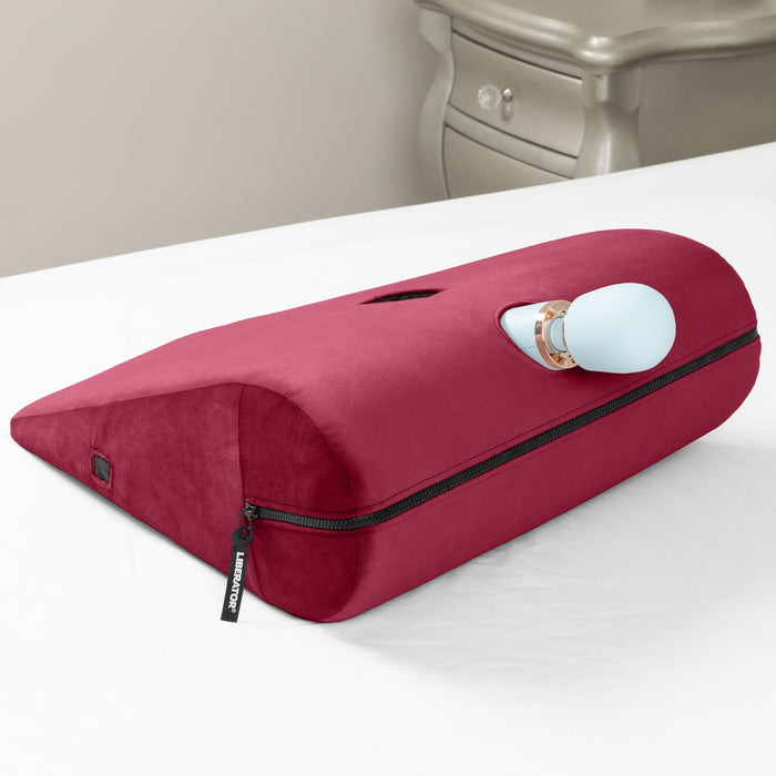 The Liberator Axis Magic Wand Hands-Free Sex Toy Mount is a red wedge-shaped ergonomic pillow with a zipper and slot that secures a blue wand massager, enabling hands-free use on a white bed near a silver nightstand.