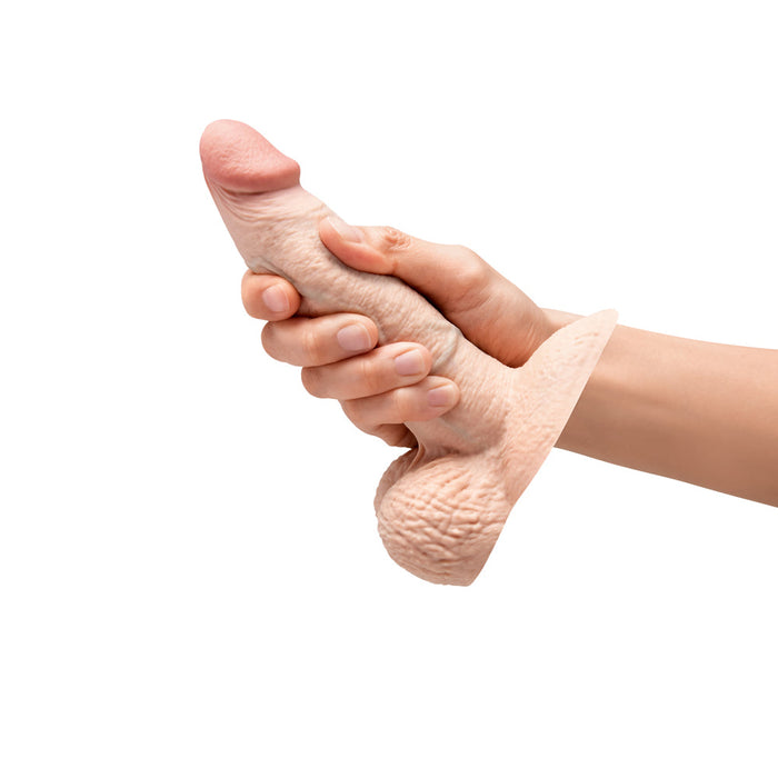 A hand holds the b-Vibe Slipskin Girthy Real Feel 6 Inch Silicone Dildo in Vanilla Tone, featuring a suction base and lifelike silicone detail for an authentic, body-safe experience against a white background.