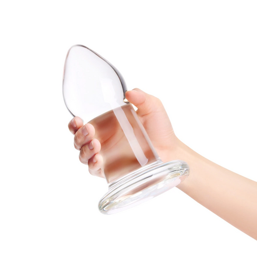 Hand holding a large, clear 7.5-inch glass anal dilator plug with a tapered head and a wide, flared base.