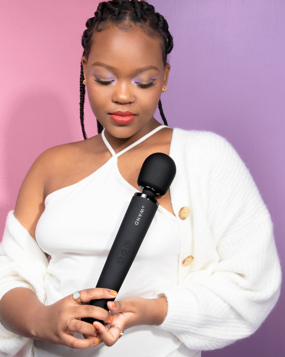 A woman with braided hair and a white outfit holds a powerful sex toy, the Le Wand Powerful Cordless Vibrator - Black, against a purple background.