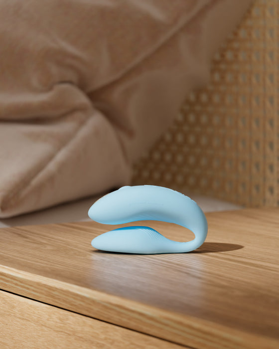 A modern, sleek blue We-Vibe Chorus Remote & App Controlled Couples' Vibrator resting on a wooden surface against a cozy, textured background.