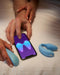 A person's hand interacting with a smartphone app to control the We-Vibe Chorus Remote & App Controlled Couples' Vibrator - Blue, featuring touch-sensitive vibrations, with other wearable technology devices placed on the bed nearby.