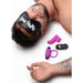 A topless man lies on a white surface wearing a black eye mask inscribed with "BANG ME!" from the BANG! Couple's Cock Ring, Finger Vibe, Bullet & Blindfold Kit - Purple by XR Brands. Beneath his arm, there are three purple and black sex toys from the kit, including a vibrating bullet. A tattoo is visible near his right wrist.