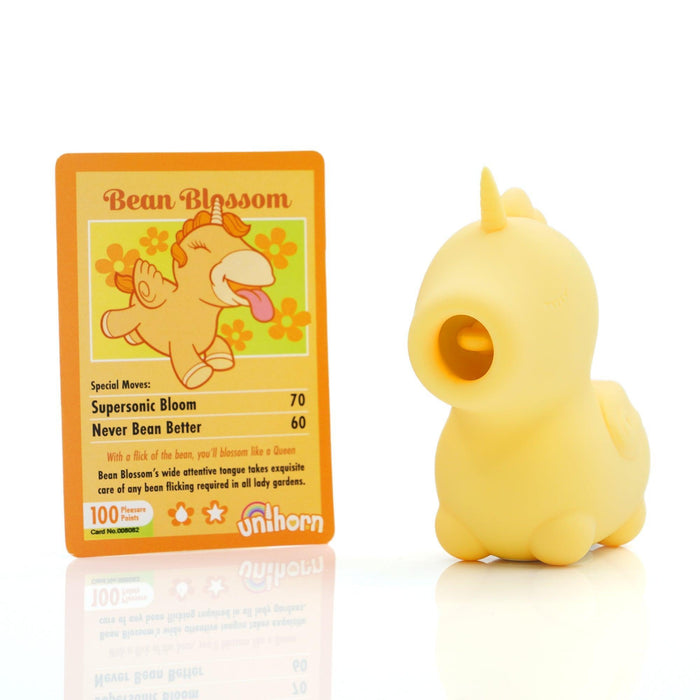 Positioned next to a vibrant card is the Bean Blossom Unicorn Shaped Licking Tongue Vibrator in yellow, crafted by Creative Conceptions. This charming toy, resembling a yellow unicorn with short legs and a round body, exudes charm and quality made from premium silicone. The accompanying card features Bean Blossom against a floral background and displays moves such as "Supersonic Bloom" and "Never Bean Better.
