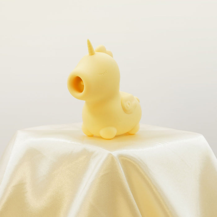 The Bean Blossom Unicorn Shaped Licking Tongue Vibrator in yellow by Creative Conceptions, a soft toy with closed eyes and a small horn, rests on a smooth, folded cream-colored cloth against a neutral background. Featuring premium silicone construction and USB rechargeable capabilities with 20 functions for endless delight.